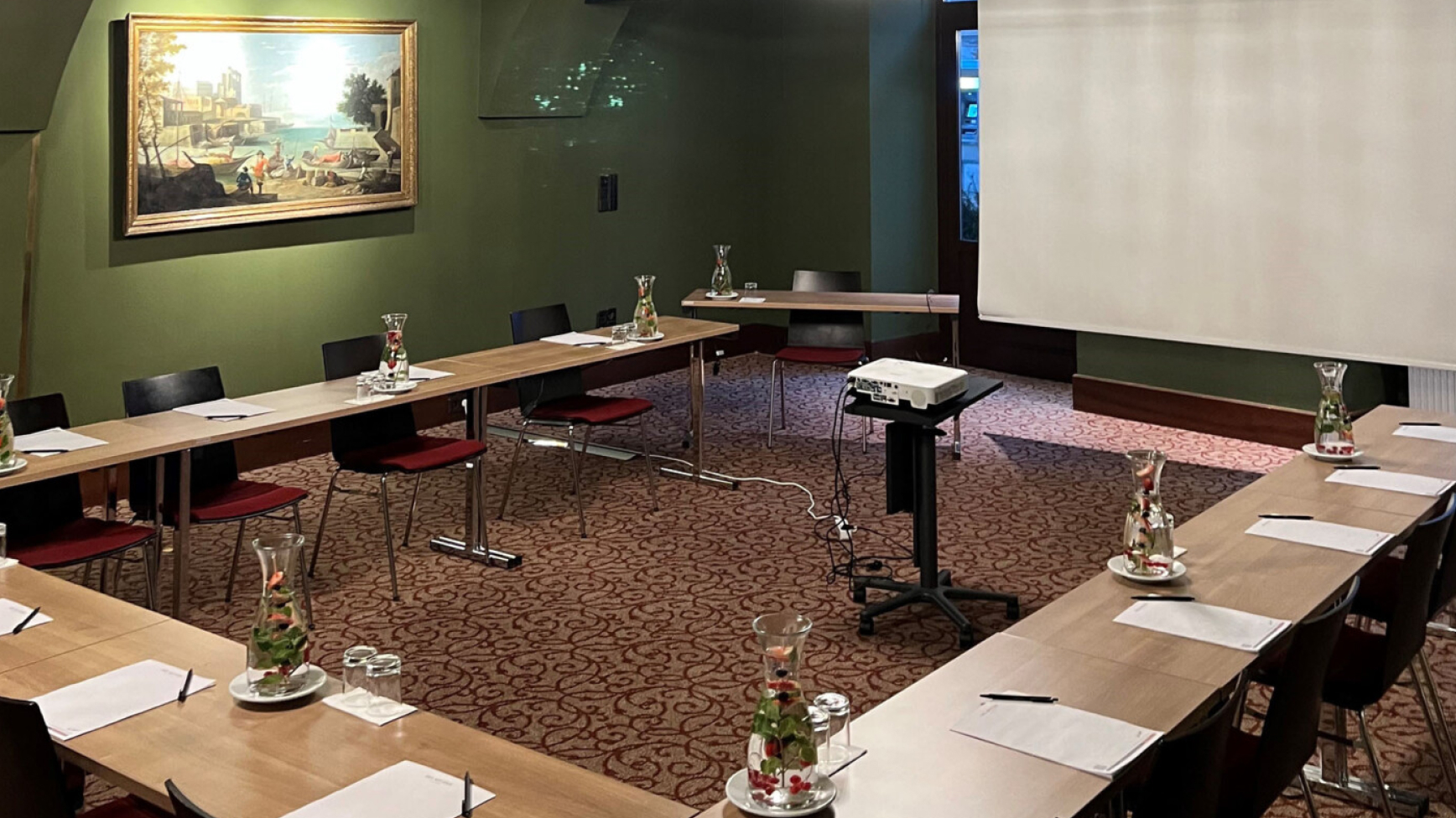 Ein Konferenzraum mit u-förmiger Tischordnung, Projektor und Leinwand. An der Wand hängt ein Gemälde.