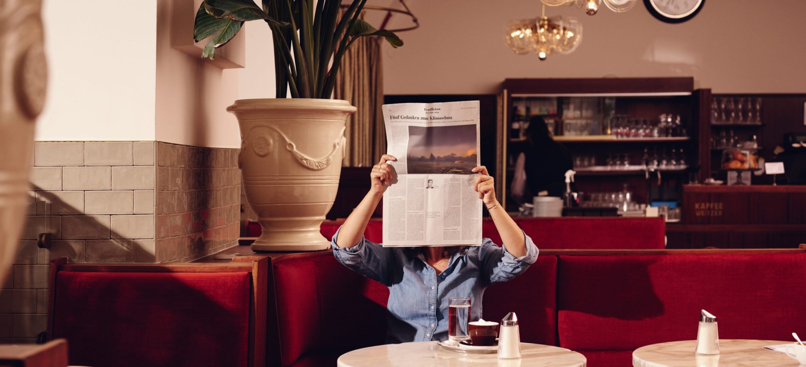 Person liest Zeitung "Tief Gefroren aus Klausurhitze" im Café mit roten Sitzen. Kaffeetasse, Glas Wasser und Zuckerstreuer.