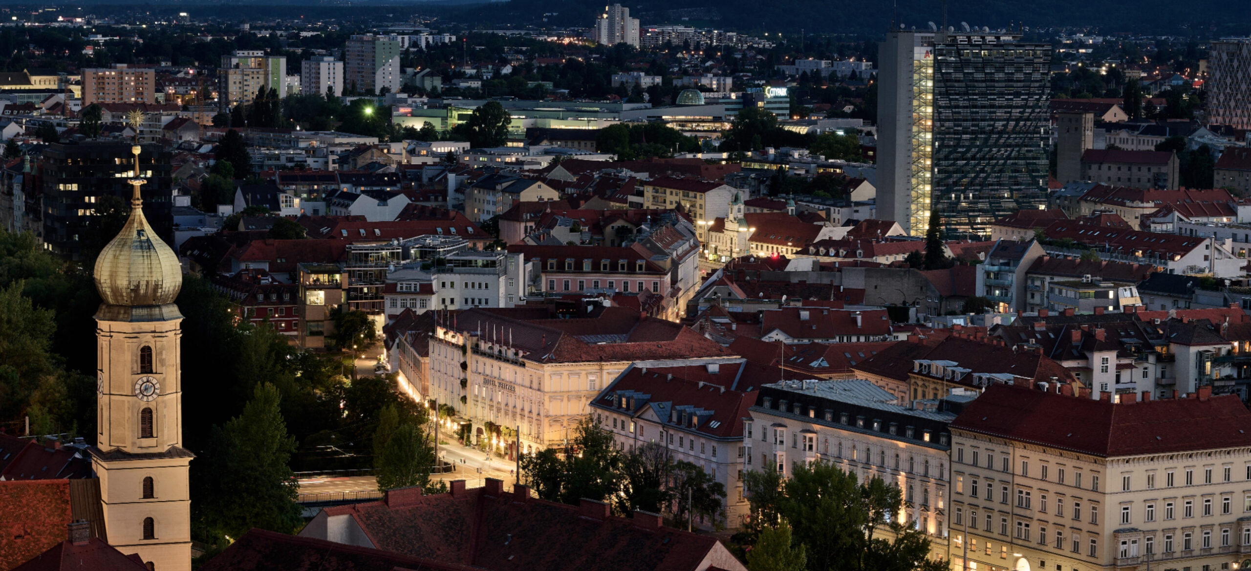 Ein Panoramablick auf Graz bei Nacht mit einem Glockenturm und roten Dächern im Vordergrund.