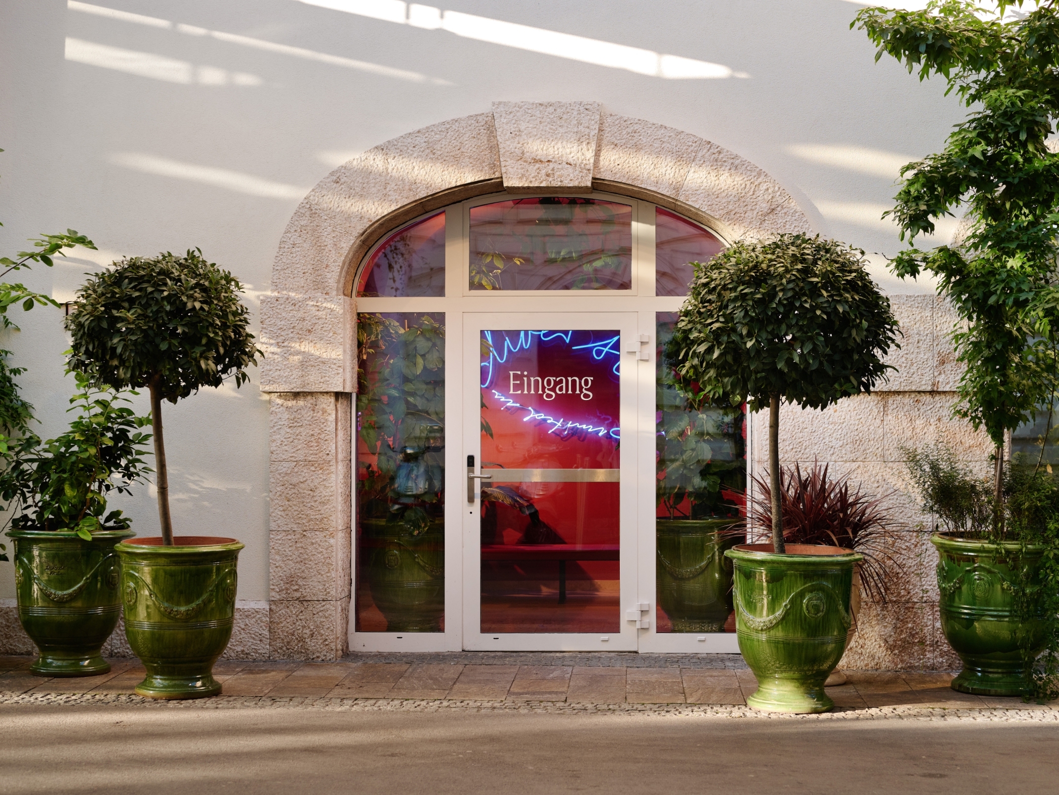 Gewölbter Eingang mit der Neonaufschrift „Eingang“ und grünen Topfpflanzen an den Seiten.