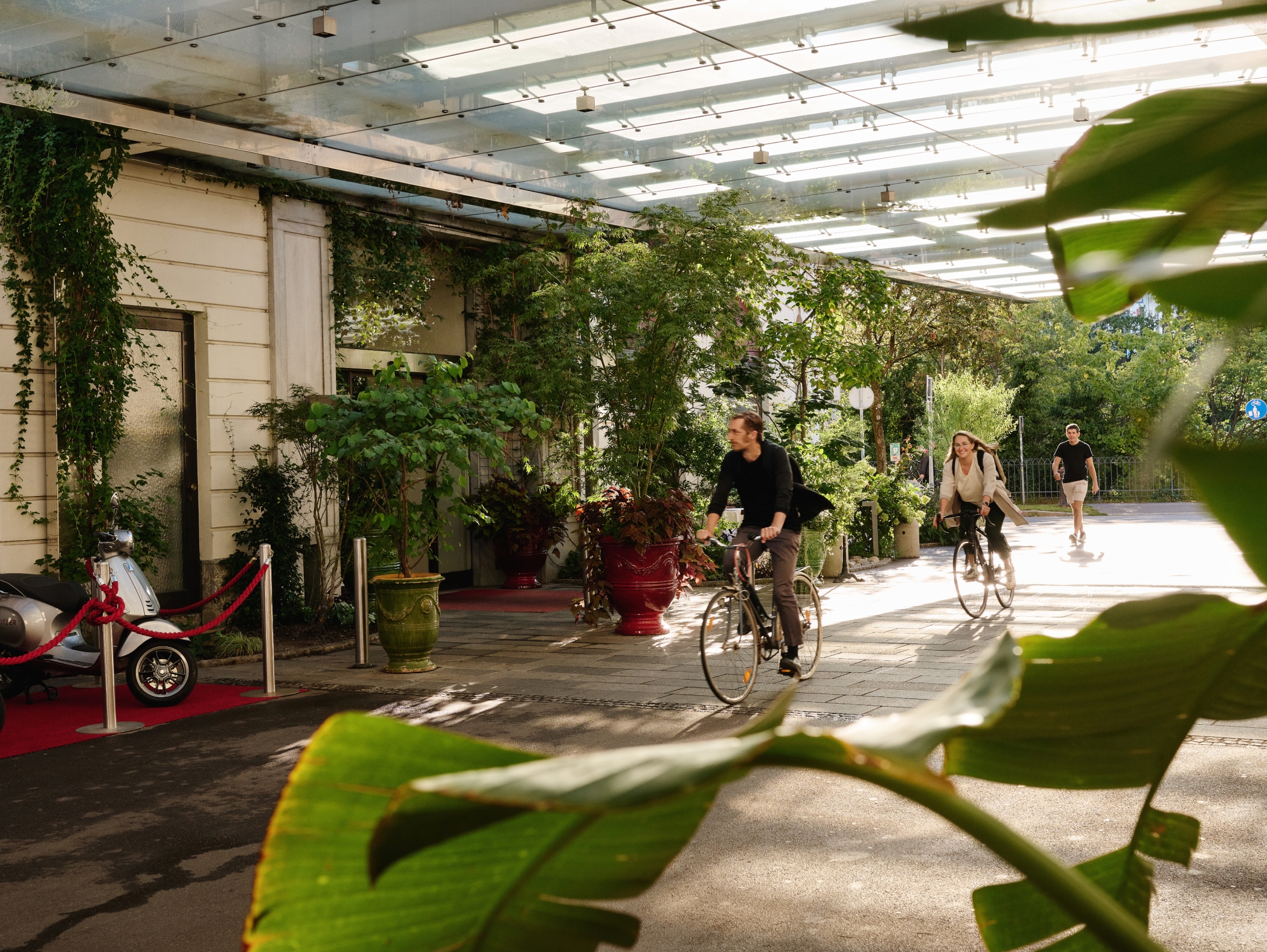 Zwei Radfahrer unter einem Glasdach, roter Teppich und Vespa in der Nähe.