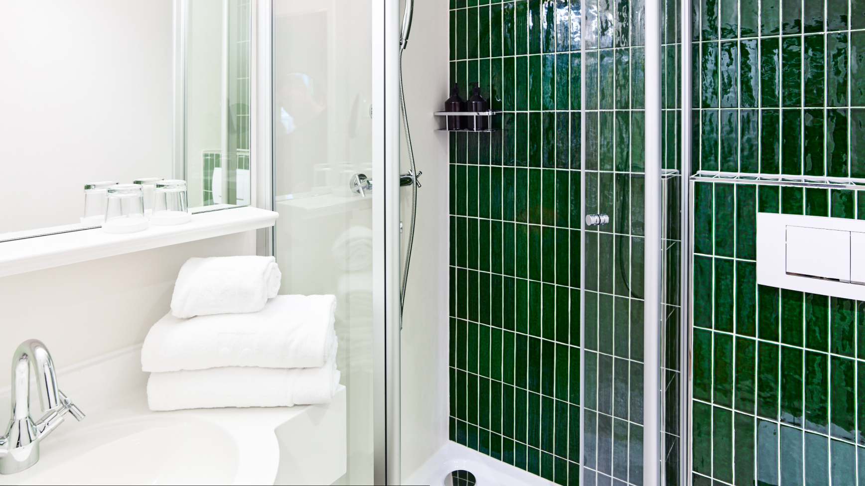 Ein Badezimmer verfügt über grüne Kacheln in der Dusche, einen Waschtisch mit Handtüchern und Gläsern sowie eine Duschtrennwand.