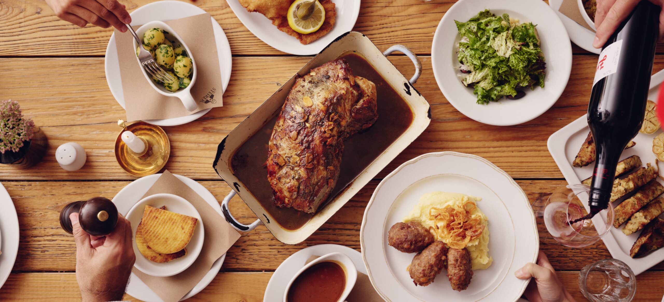 Draufsicht auf Esstisch mit Braten, Beilagen, Salat und Wein, Hände im Bild.