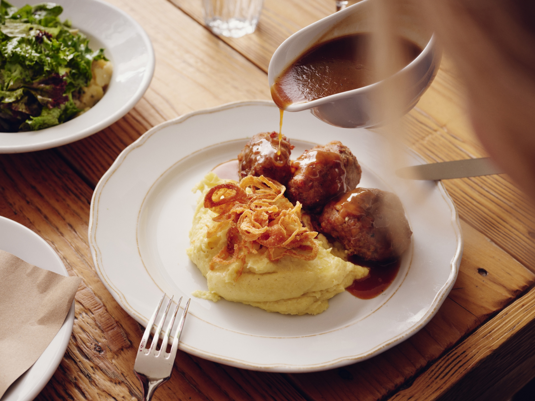 Ein Teller mit Kartoffelpüree, Fleischbällchen, Zwiebelringen und Soße wird auf einem Holztisch serviert.