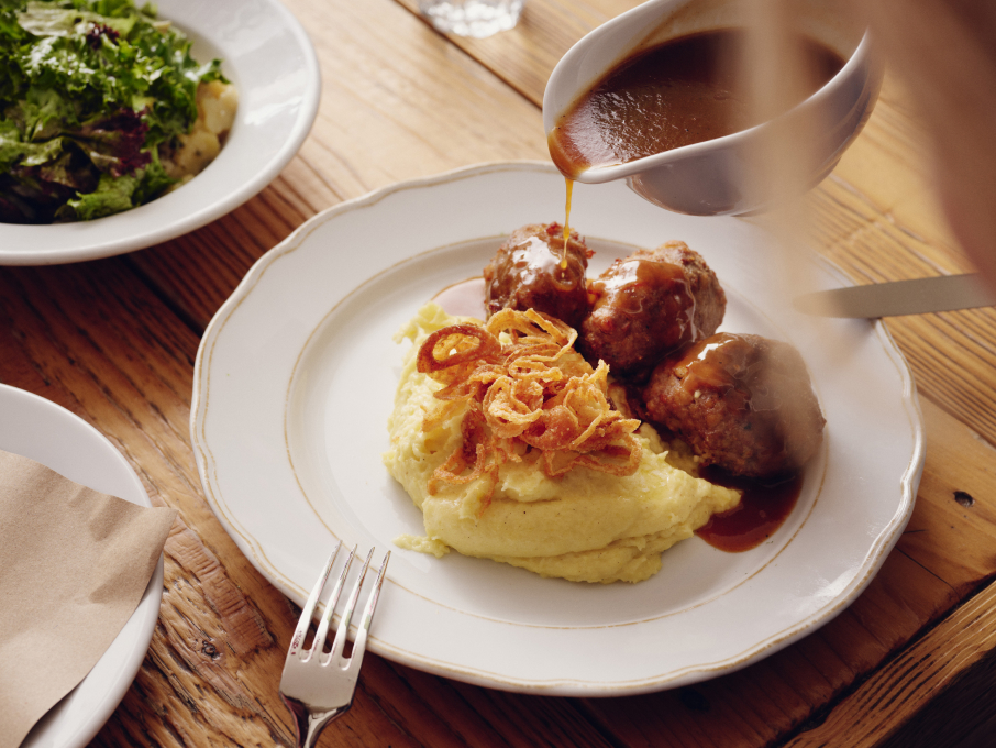 Teller mit Fleischbällchen, Kartoffelpüree und Zwiebeln; Soße wird über das Essen gegossen.