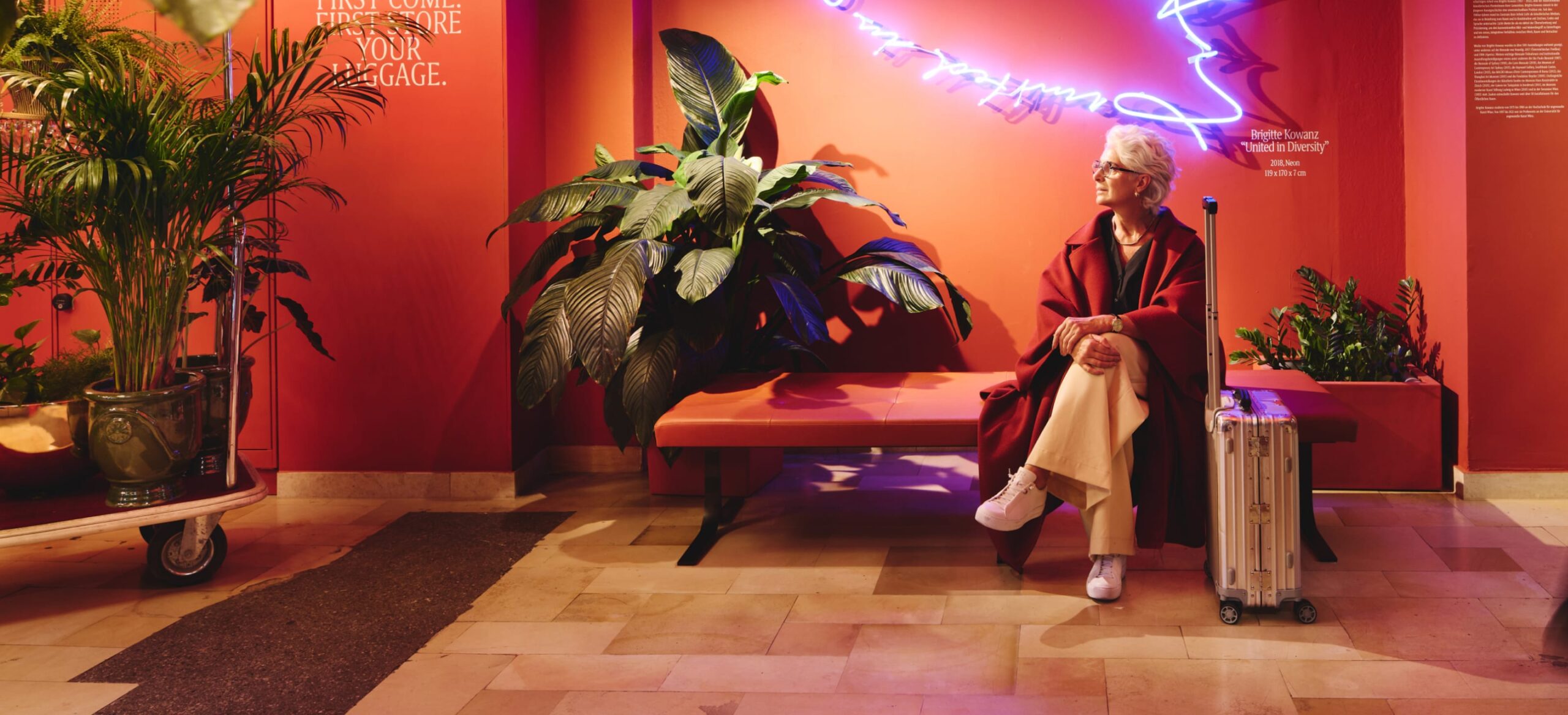 Eine Frau sitzt mit Gepäck in der „Brighter Lounge“ unter der Neonarbeit „United in Diversity“.