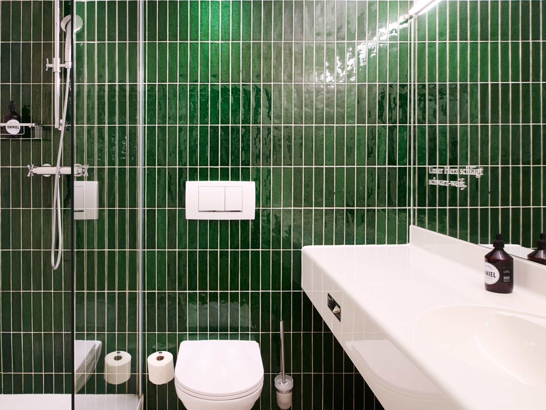 Badezimmer mit grünen Kacheln, Toilette, Waschbecken und der Aufschrift "Unser Herz schlägt schwarz-weiß."