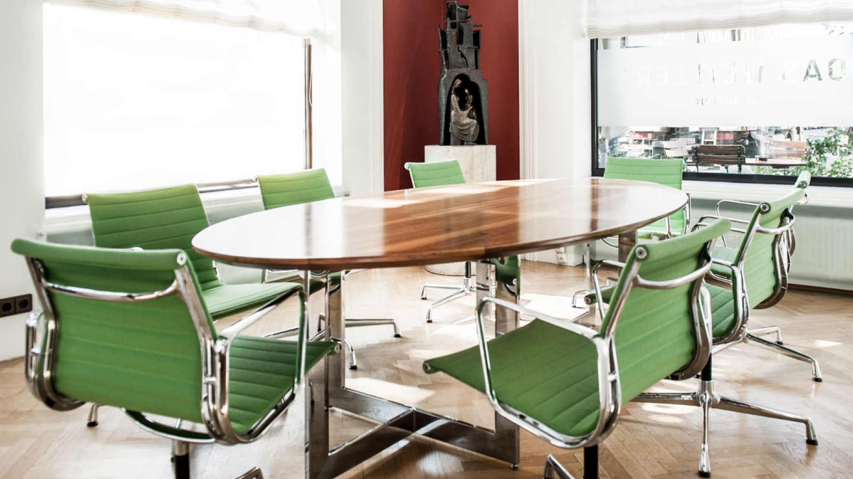 Konferenzraum mit ovalem Holztisch und grünen Stühlen, Skulptur im Hintergrund.