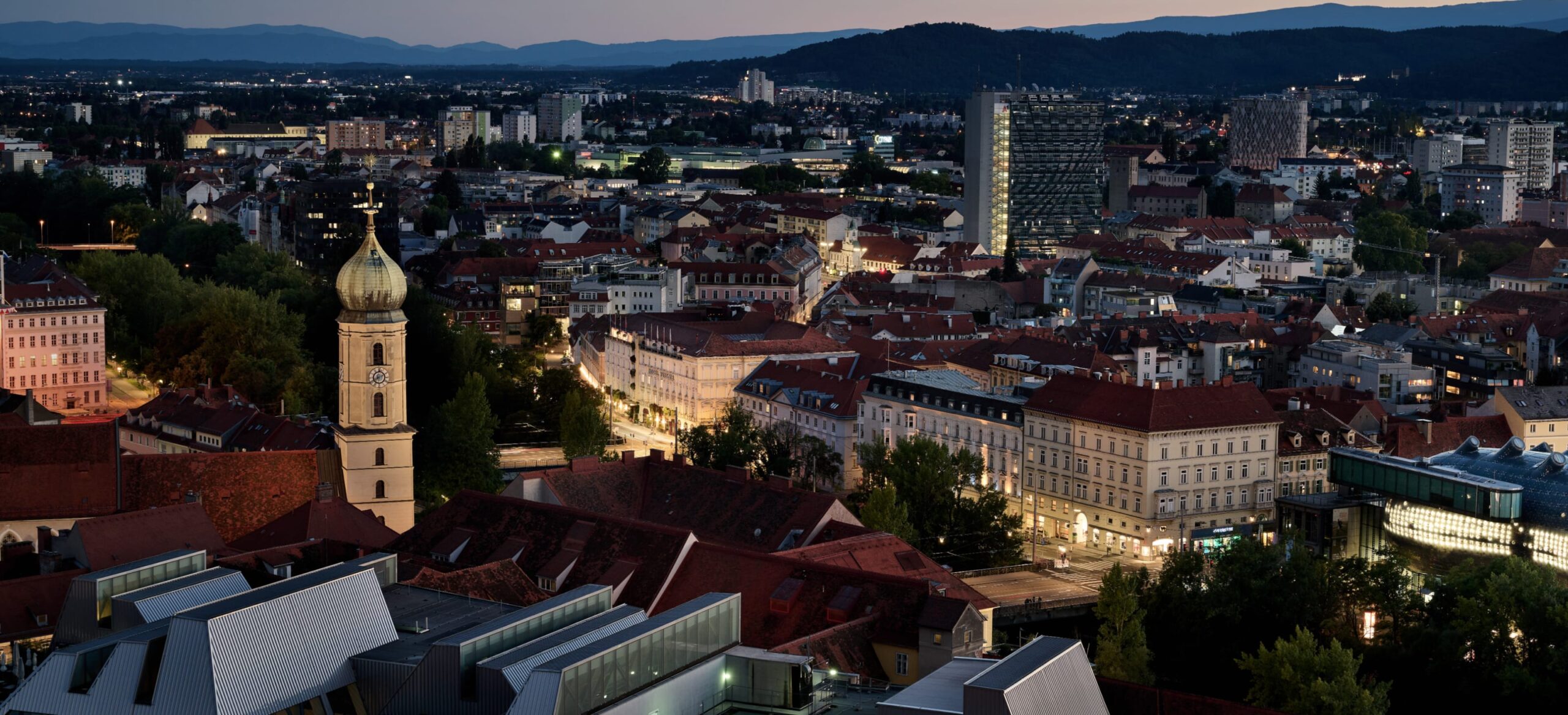 Eine weite Aufnahme von Graz, Österreich, bei Nacht. Im Vordergrund befindet sich ein Kirchturm.