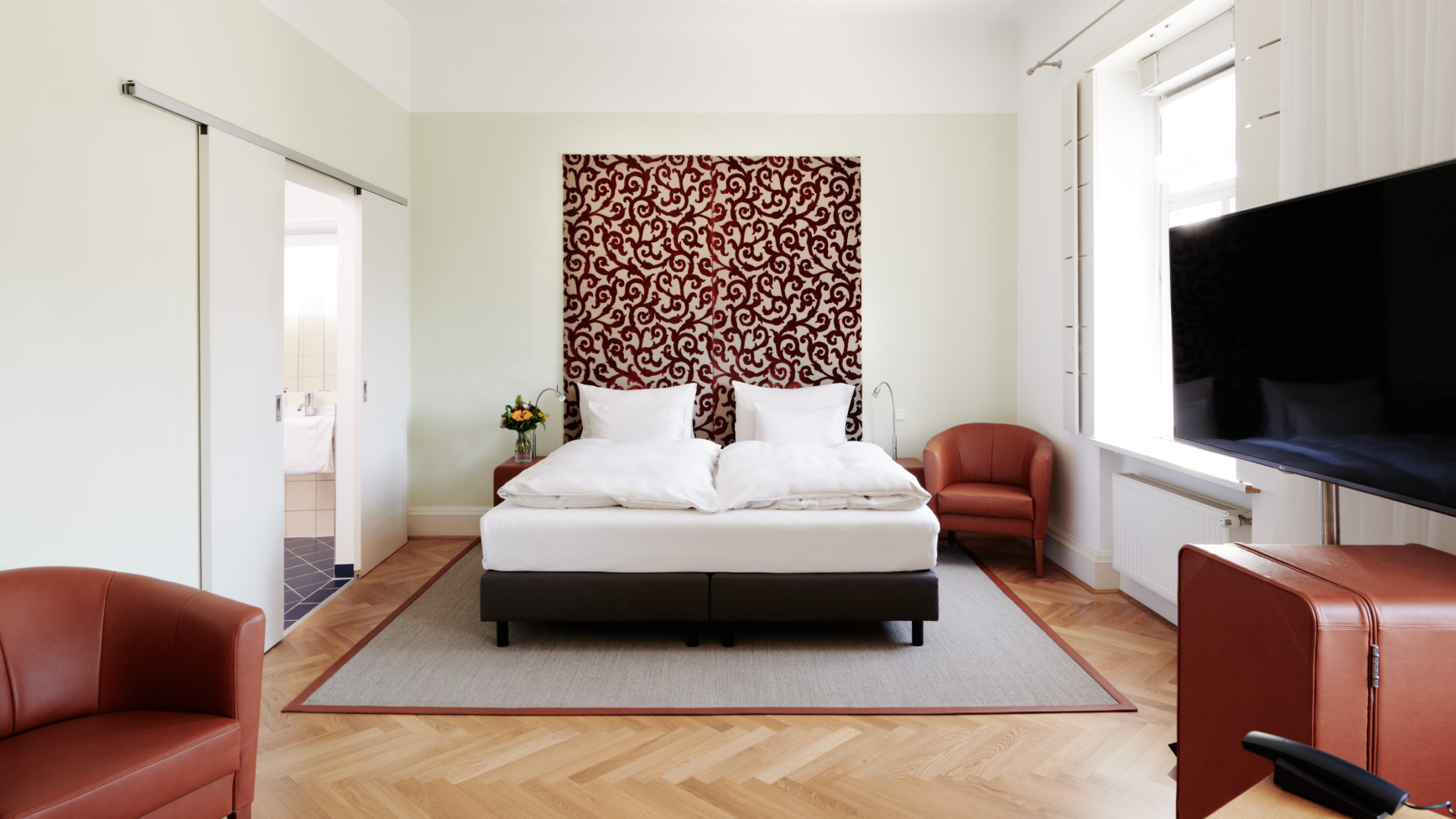 Ein Schlafzimmer mit einem Bett, zwei Stühlen und einem Fernseher. Das Zimmer hat Holzböden und eine helle Farbpalette.