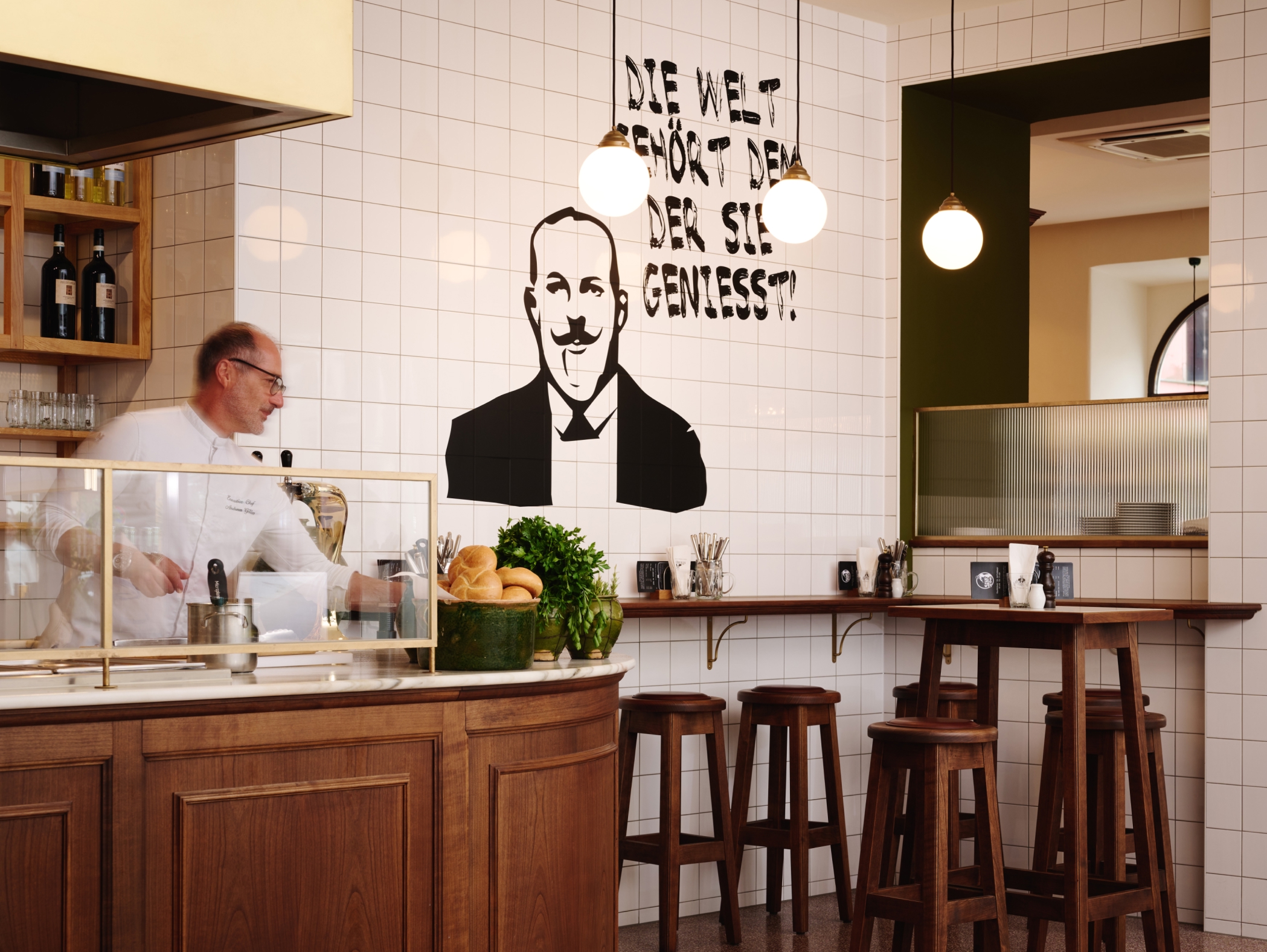 Innenraum mit Koch, Barhockern und Wandbild: "Die Welt gehört dem, der sie genießt!".