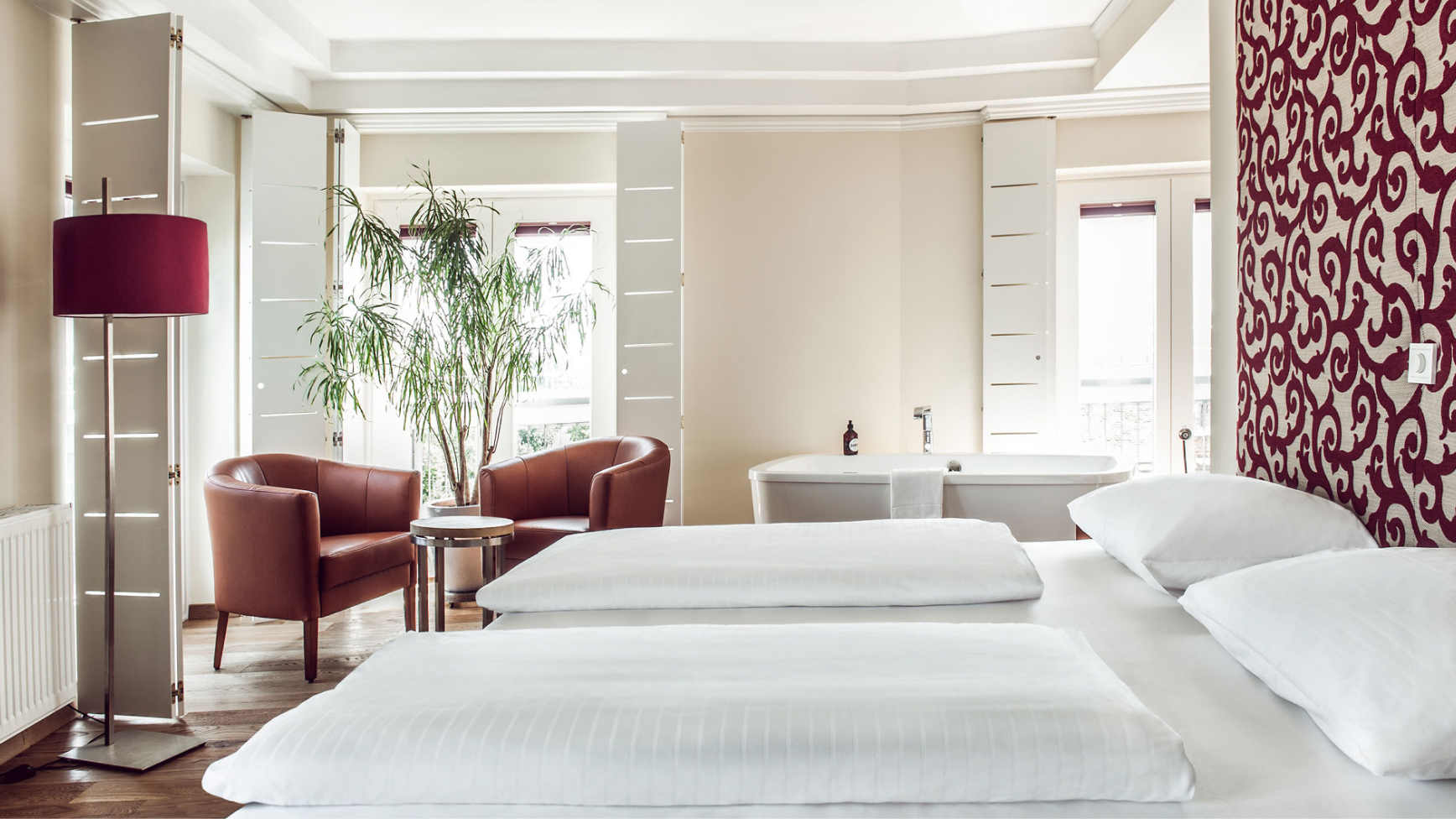 Ein helles Hotelzimmer mit Sesseln, Zimmerpflanze, Badewanne und großem Bett mit weißer Bettwäsche.