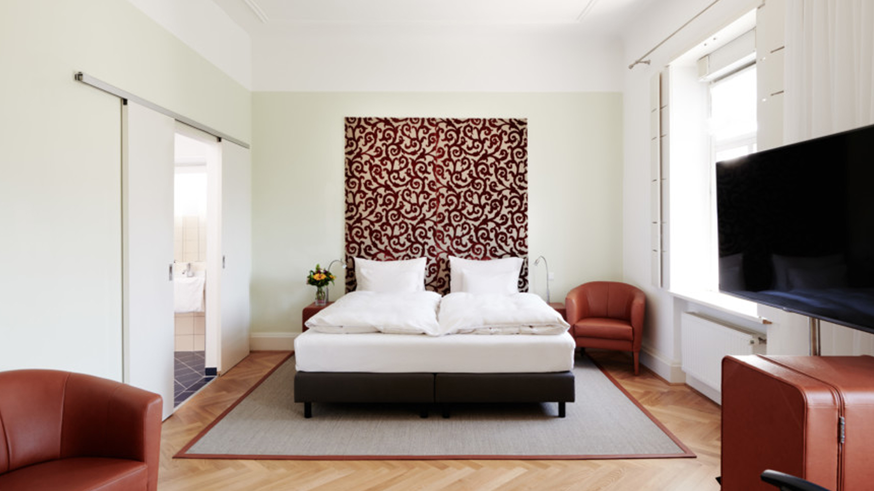 Ein helles Schlafzimmer mit einem Doppelbett, zwei Sesseln und einem Fernseher.