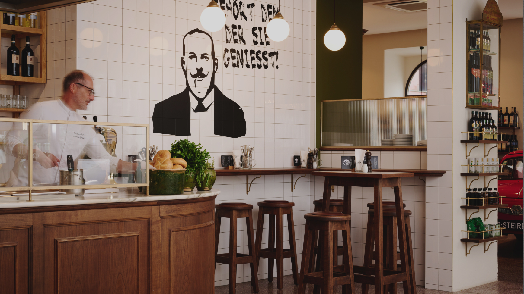 Innenansicht von Bäckerei mit Koch, Barhockern, Weinregalen und Aufschrift "Gehört dem, der sie geniesst!".