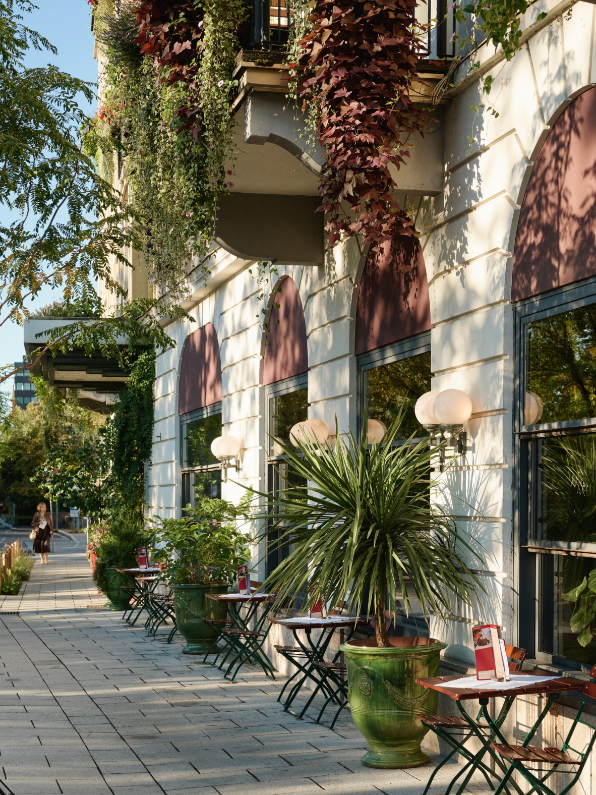 Außenaufnahme eines Cafés mit Tischen und Stühlen auf dem Gehweg. Fassade mit Pflanzen und Fenstern.
