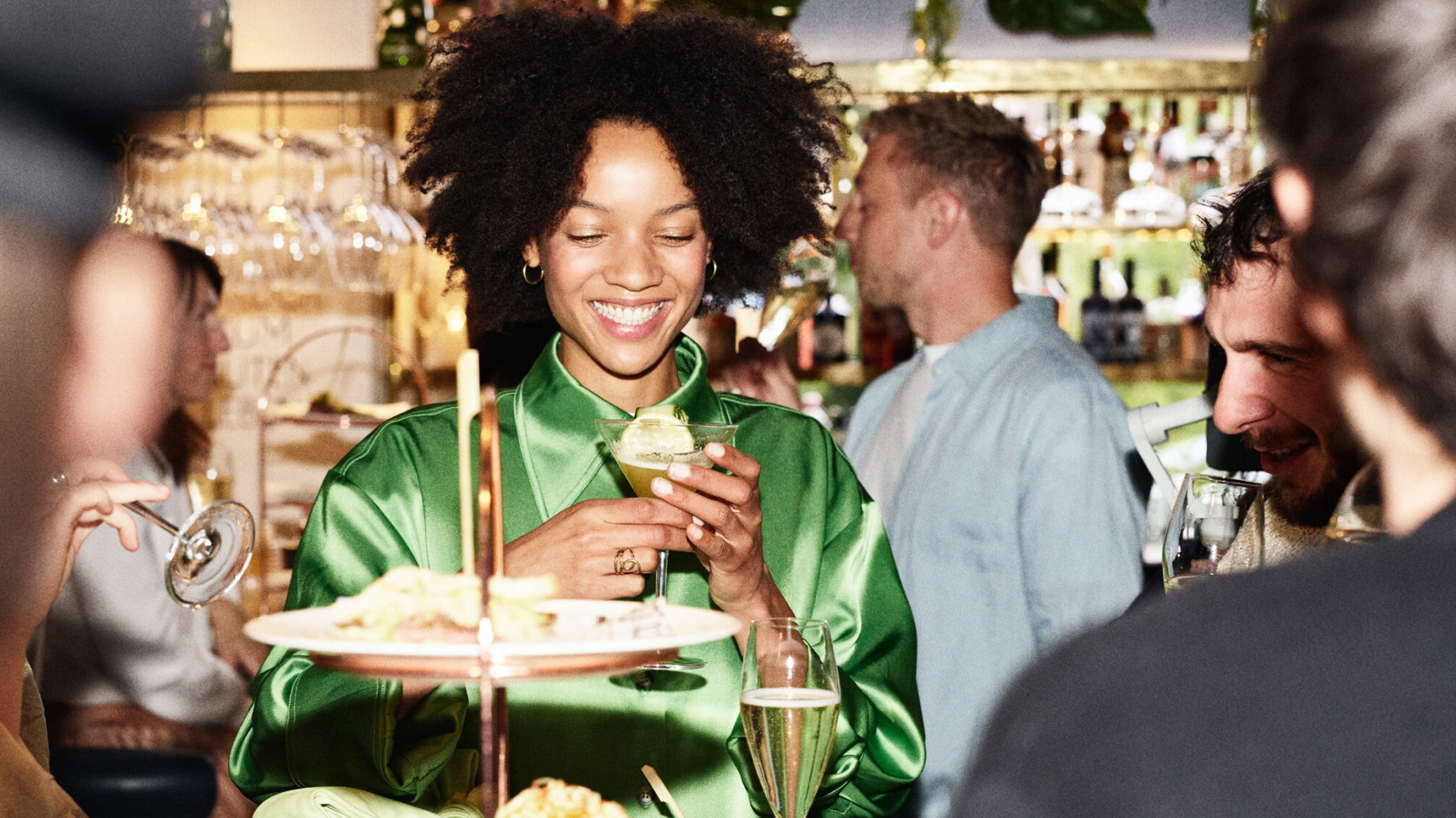 Lachende Frau im Grünen mit Getränk, umgeben von Gästen in einer Bar.