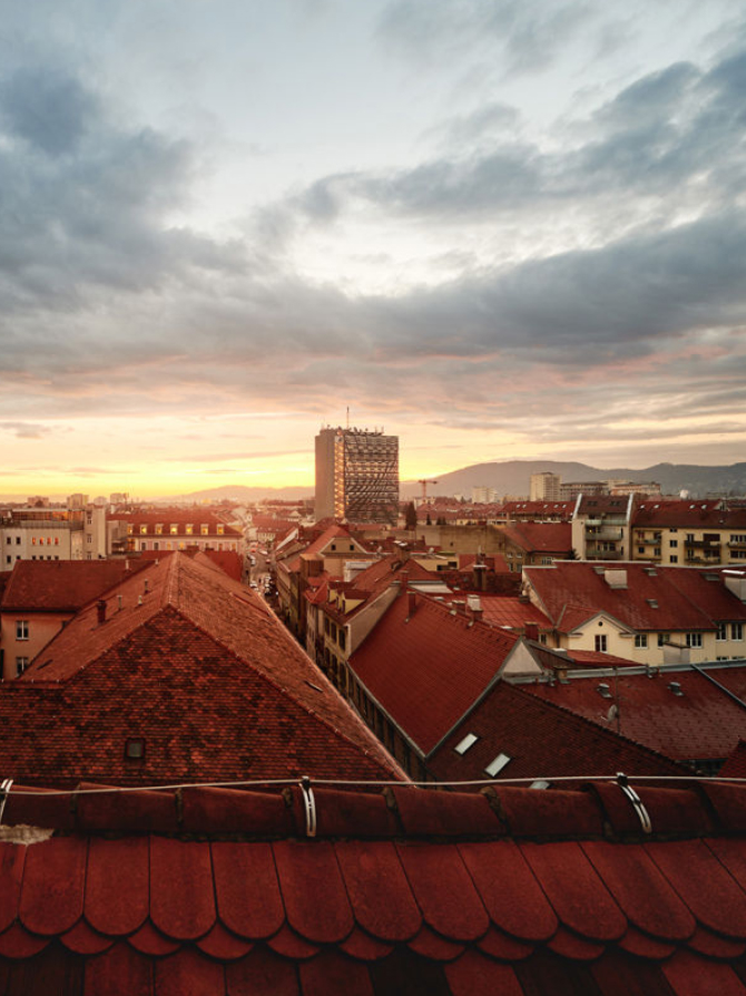 Luftaufnahme von Graz, Österreich, mit roten Dächern, einem hohen Gebäude und Bergen im Hintergrund.