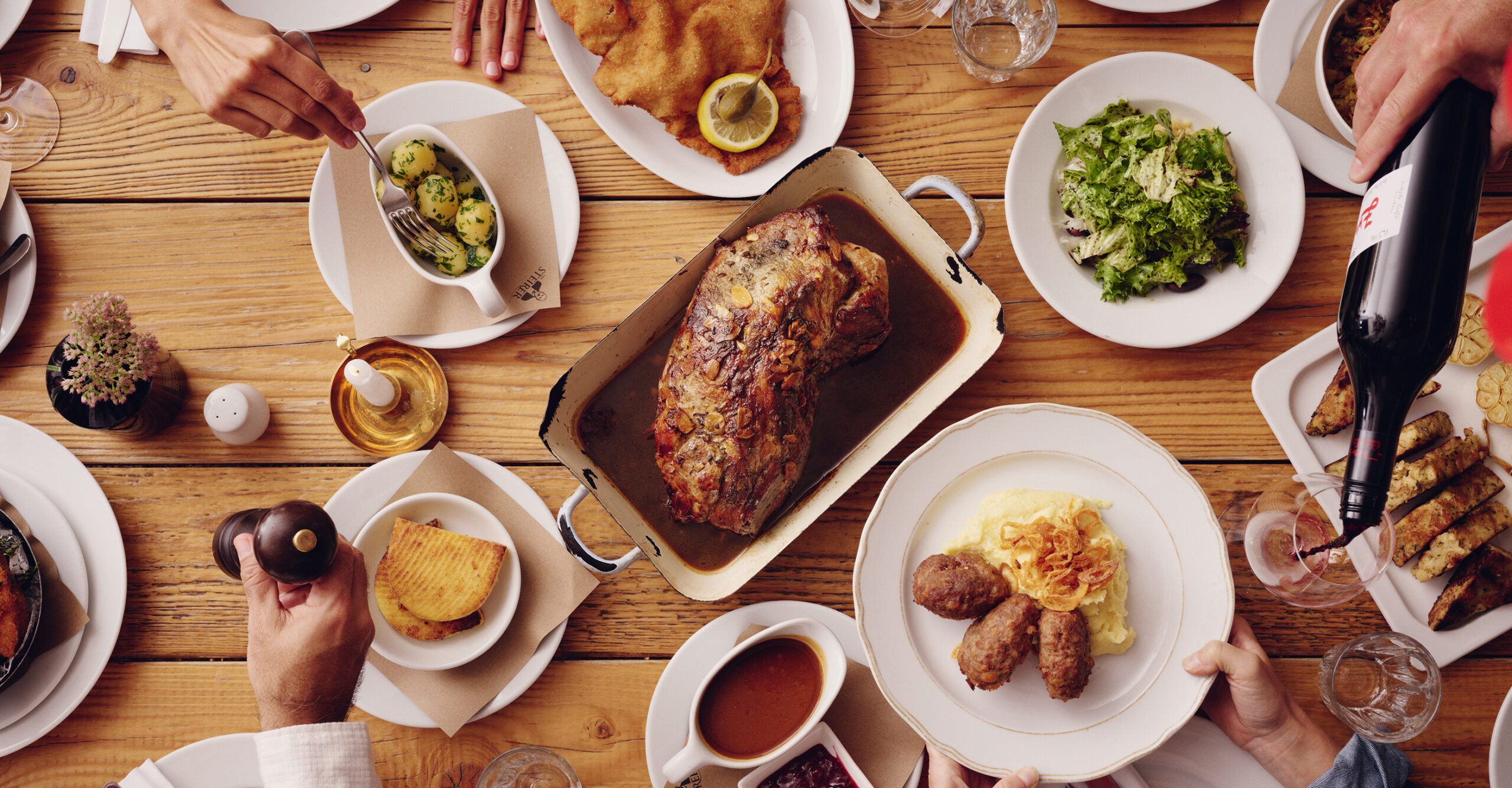 Draufsicht auf festliches Essen: Braten, Salat, Knödel, Schnitzel, Wein. Hände und Teller.