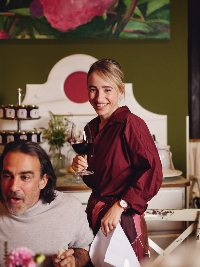 Eine lachende Frau mit dunkler Burgunderbluse hält ein Glas Rotwein in einem Esszimmer.