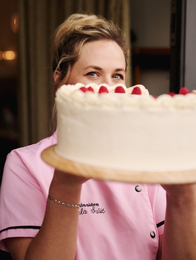 "La Sušić"-Bäckerin hält einen Himbeerkuchen hoch, lächelt über den Kuchen.