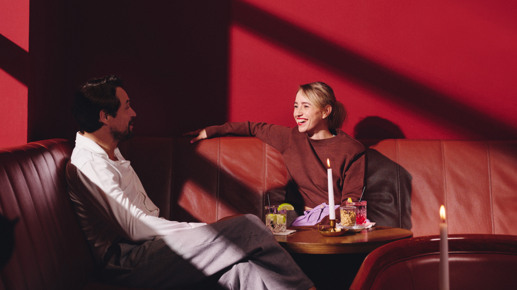 Ein Mann und eine Frau sitzen in einer roten Bar auf einer Bank mit Cocktails und Kerzen.