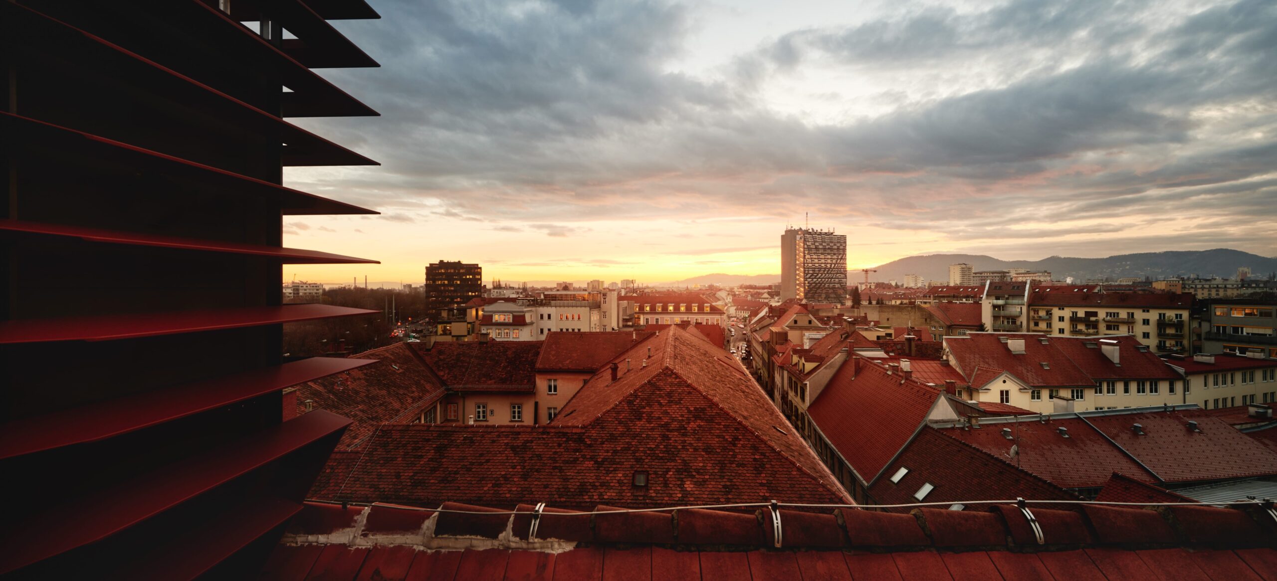 Skyline view of Graz with red roofs and a cloudy sunset sky, partially obscured by red blinds.