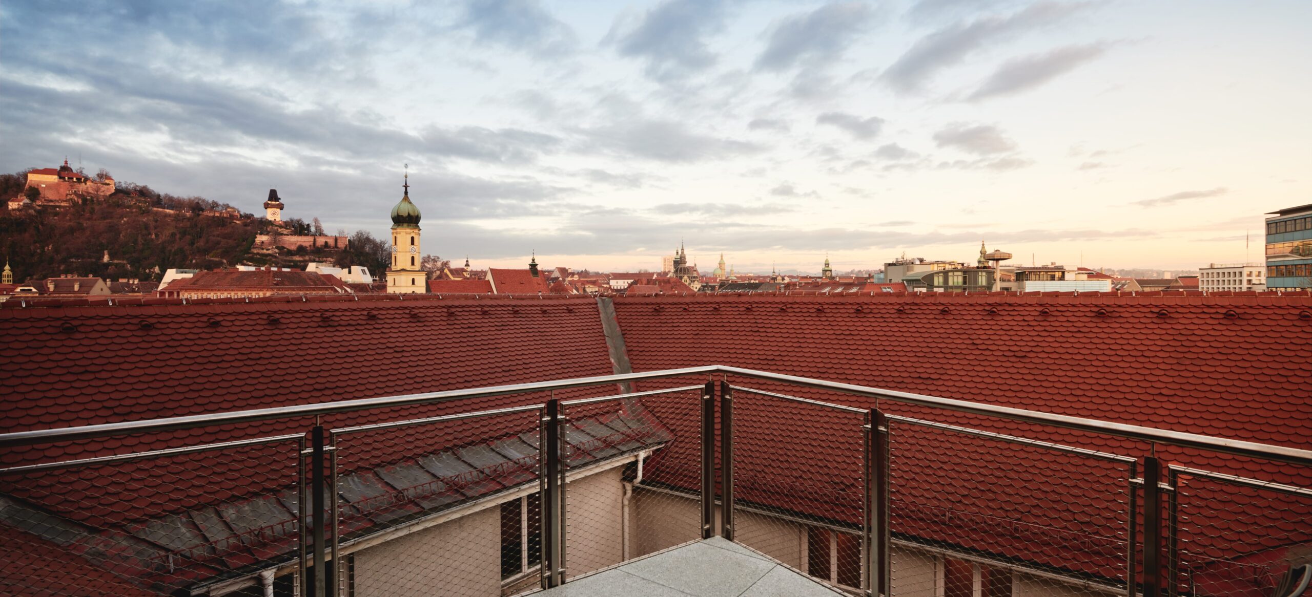 View of Graz from a balcony, red roofs, Schlossberg and clock tower under cloudy sky.