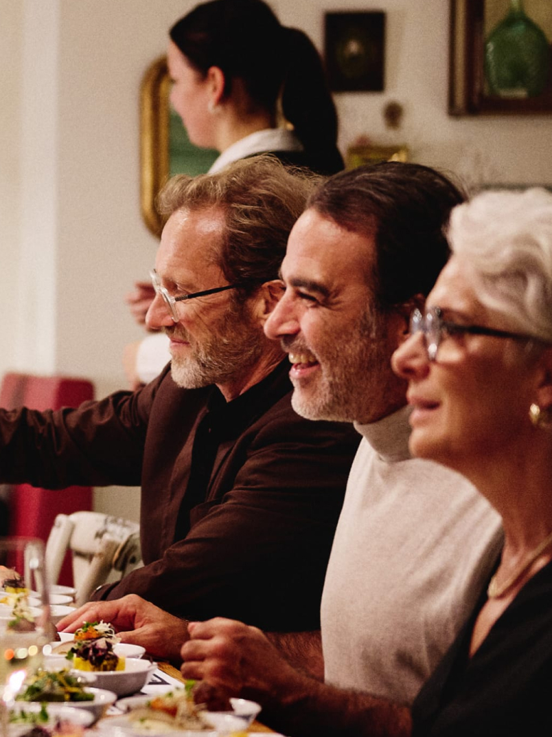 Three older people sit at a dining table, looking right, with food dishes and a server in the background.