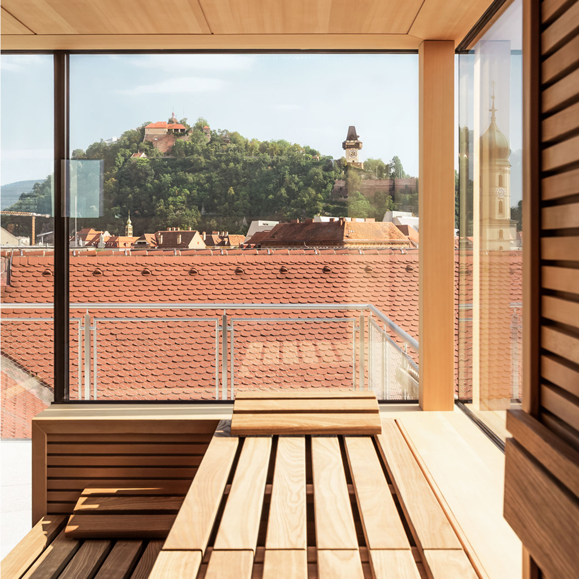 View from a wood sauna onto red rooftops in Graz, Austria, with Schlossberg and clock tower views.