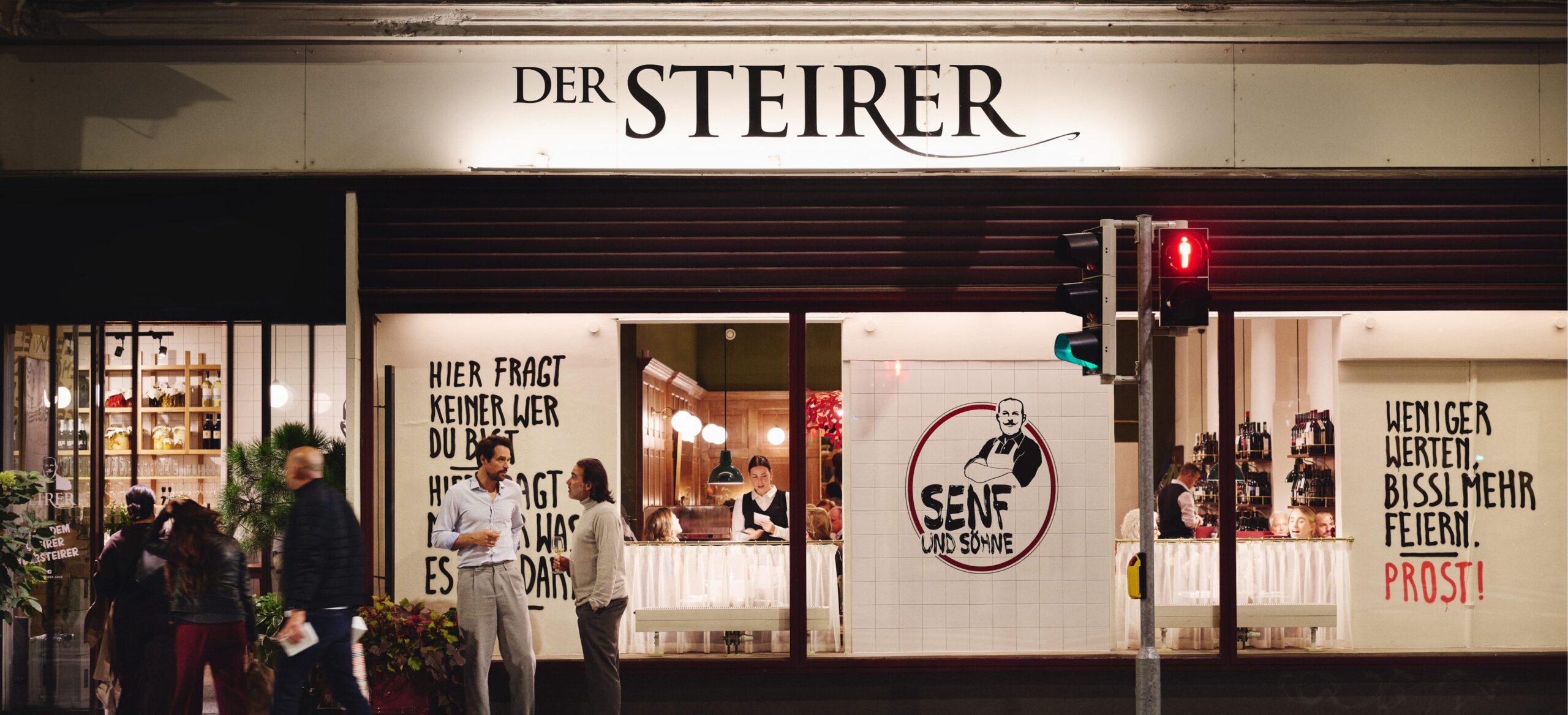 "Der Steirer" storefront with "Senf und Söhne" logo. People nearby, traffic light is red. Slogans visible on windows.
