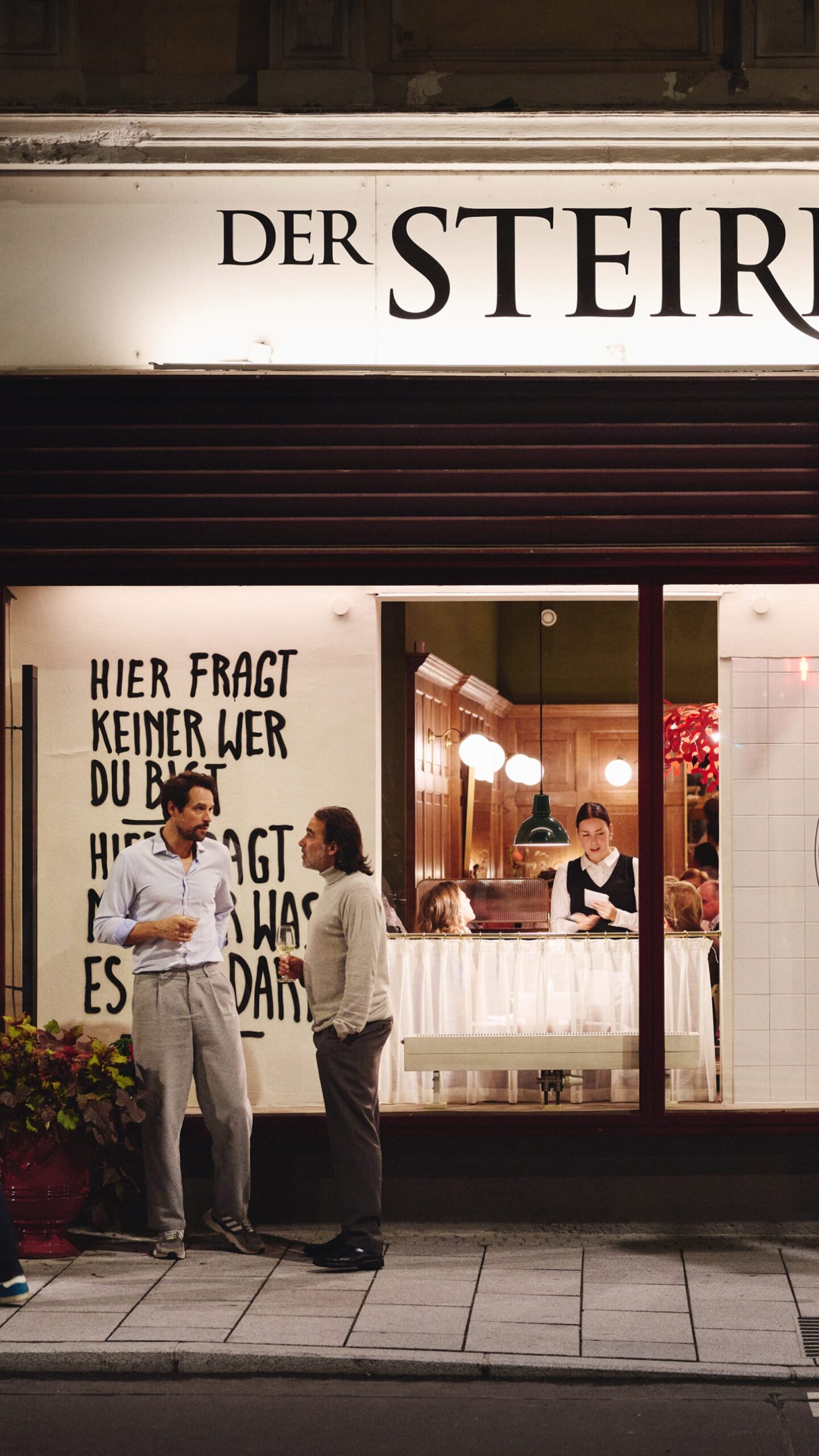 Two men converse outside "Der Steirer," sign reads "Hier fragt keiner wer du bist." A waiter is visible inside.
