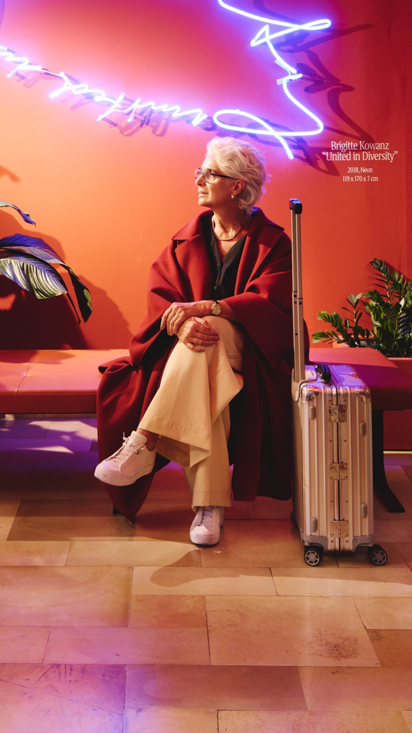 A woman with a suitcase sits in a room with Brigitte Kowanz neon art. The art reads "United in Diversity."