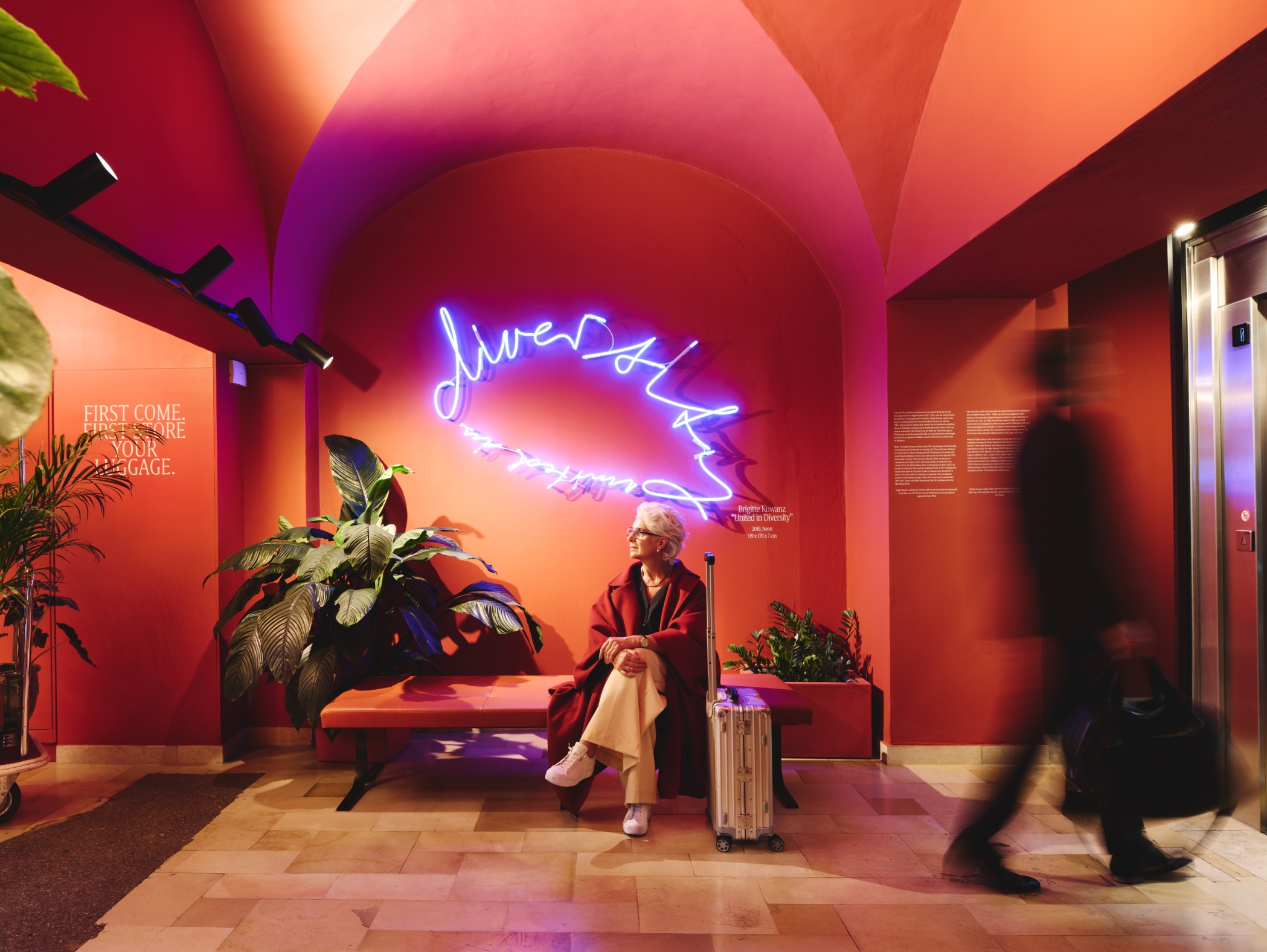 Frau wartet mit Koffer auf Bank. Neon-Text „diversity“. Person geht am Aufzug vorbei. Roter Raum.