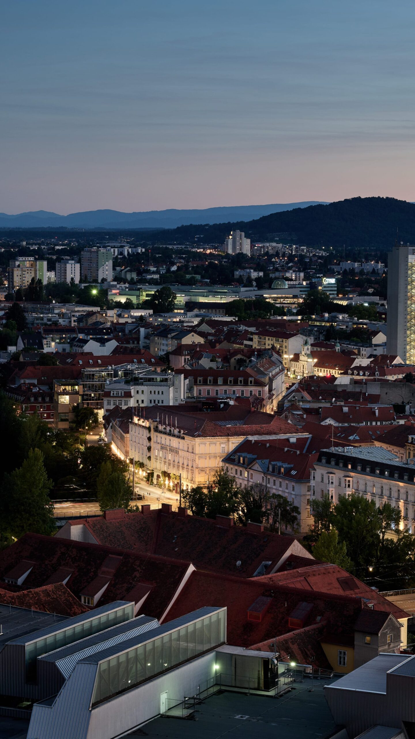 Night aerial of Graz, Austria, showing red roofs, city details, and mountains in the distance.