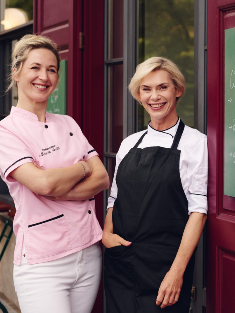 Two smiling chefs: one in a pink "Patissière Marela Sele" uniform, the other in black apron, stand near a red door.