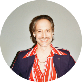 Ein Mann mit einem gestreiften Hemd, einer Goldkette und einem Blazer, der lächelt.