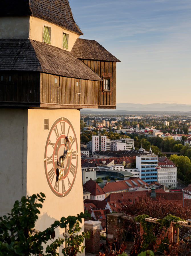 The Grazer Uhrturm clock tower with a cityscape backdrop. The clock has large Roman numerals.