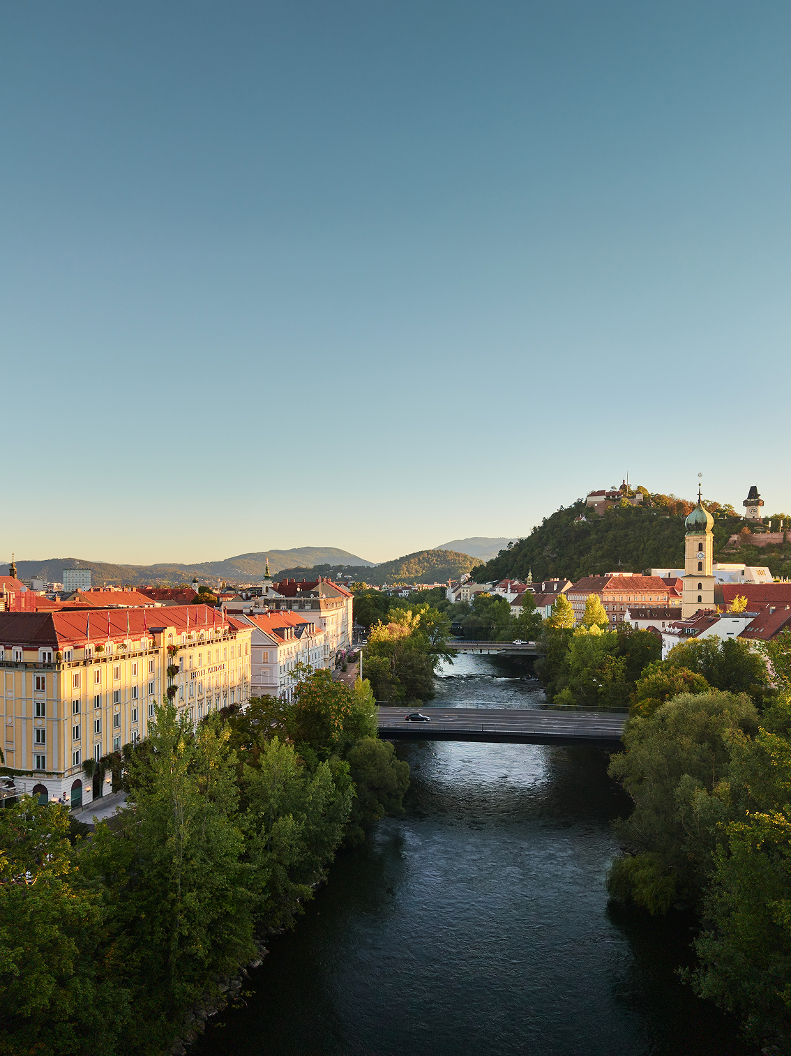 A scenic view of Graz, showcasing the Mur river, Hotel Weitzer, and the Grazer Schlossberg.