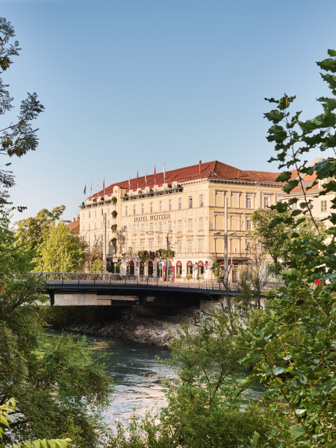View of Hotel Weitzer in Graz over a river with a bridge, surrounded by greenery.
